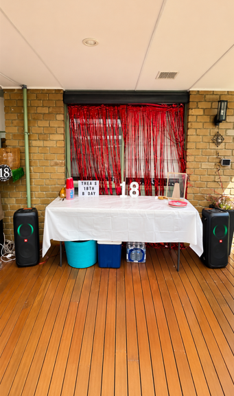 Patio birthday setup with speakers flanking a red backdrop and party table