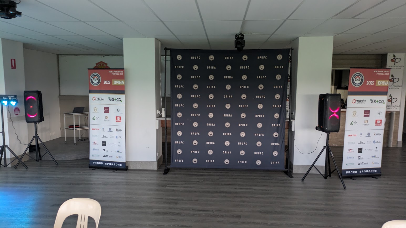 Speakers on stands flanking a sponsor backdrop at a football club function