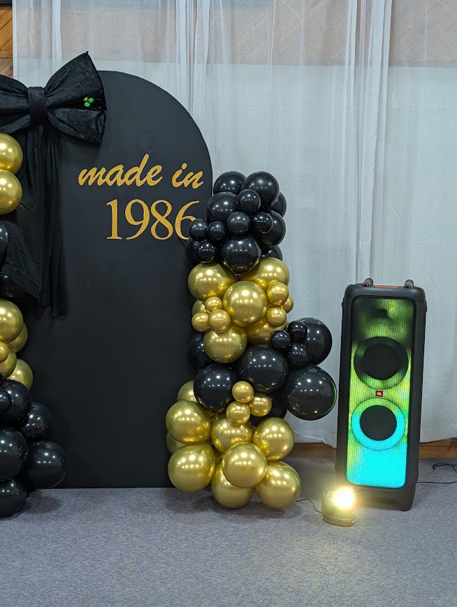 Party speaker beside a black and gold balloon garland birthday arch